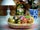 Panier décoratif rempli d’œufs de Pâques en bois peints à la main style russe Zhostovo, motifs floraux colorés, posé sur une table avec lampe, plante et horloge en arrière-plan chaleureux